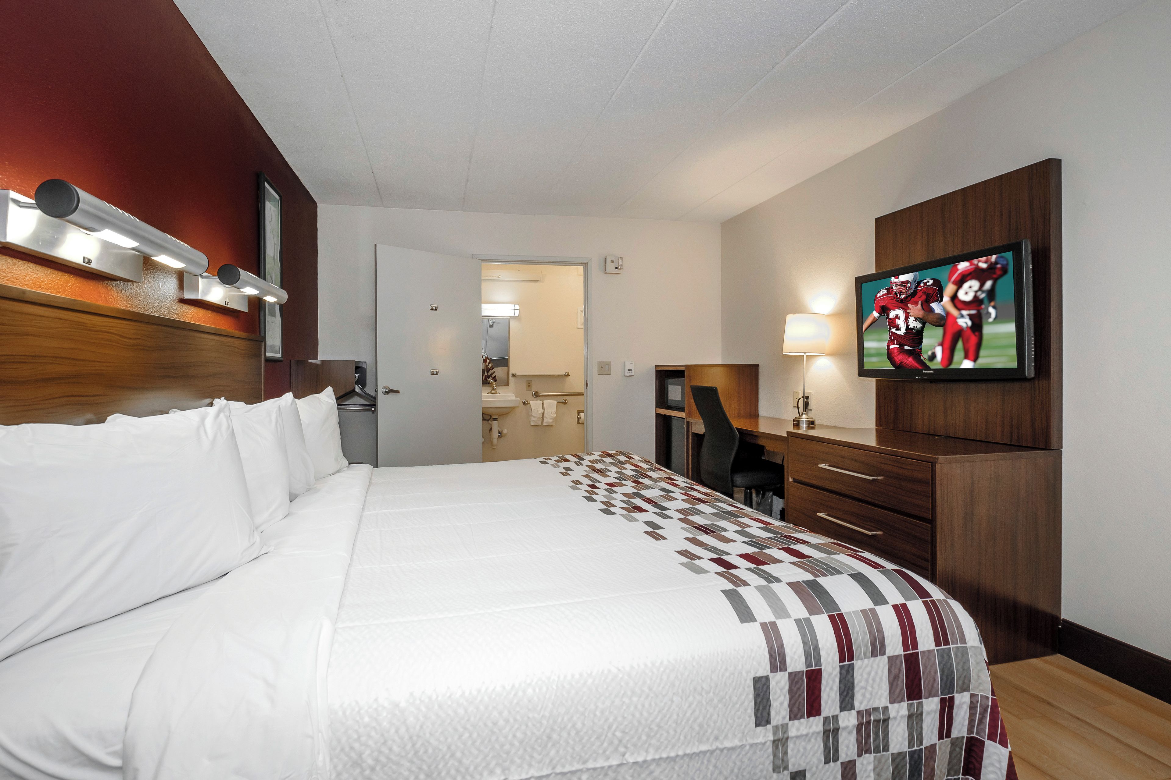 A hotel room with a large bed, a desk, a chair, a TV, and a bathroom visible through an open door.