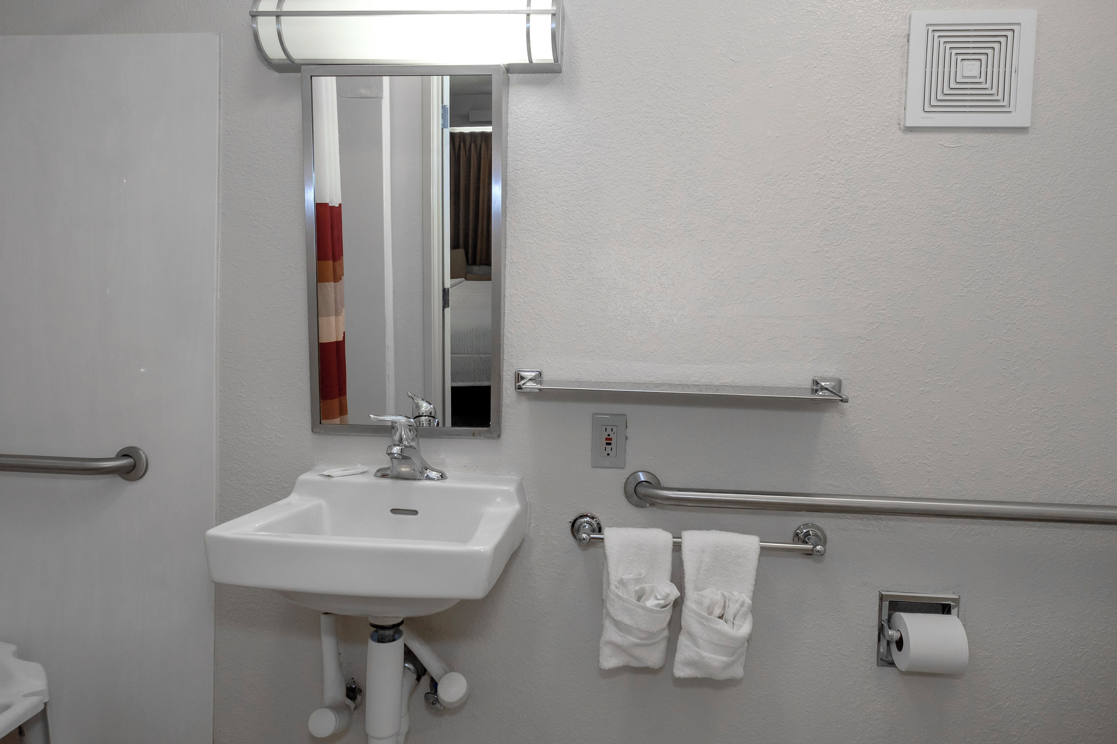 Un baño con lavamanos blanco, espejo, toallero y dispensador de papel higiénico.
