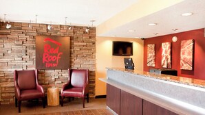 A Red Roof Inn reception area with a stone wall backdrop, two leather chairs, a wooden stool, a flat-screen TV, and artwork on the wall.