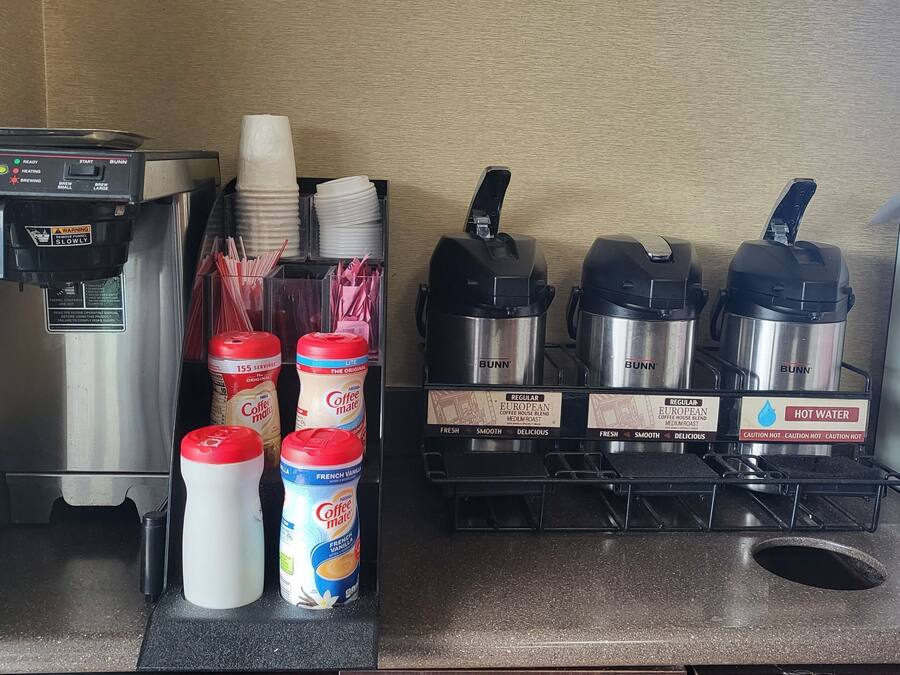 Uma estação de café com uma cafeteira Bunn, copos de papel, canudos e diversos recipientes de café, incluindo leite cremoso sabor baunilha francesa.