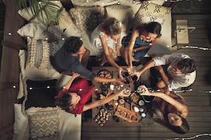 A group of people seated around a wooden table with food and drinks, in a cozy, rustic setting with wooden furniture and string lights.