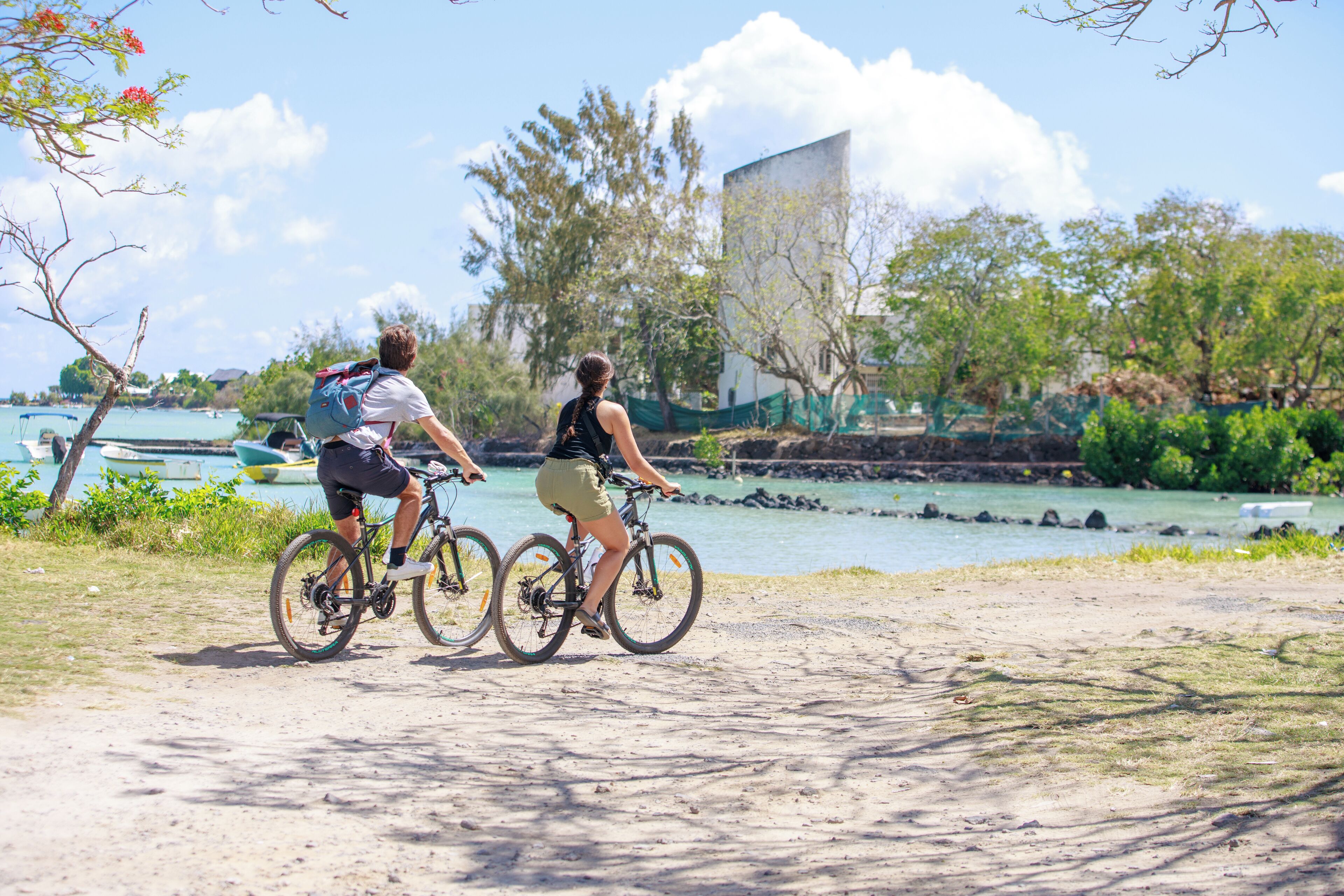 Two people on bicycles on a dirt path near a body of water with boats in the background.