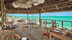 An outdoor dining area with wooden tables and chairs, thatched roof, and a view of the ocean. - Paradise Cove Boutique Hotel - Adults only (Cap Malheureux)