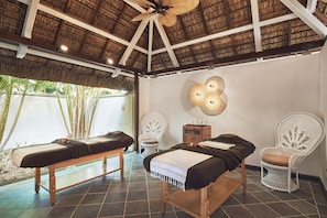 A room with two massage tables, white wicker chairs, a ceiling fan, and a thatched roof.