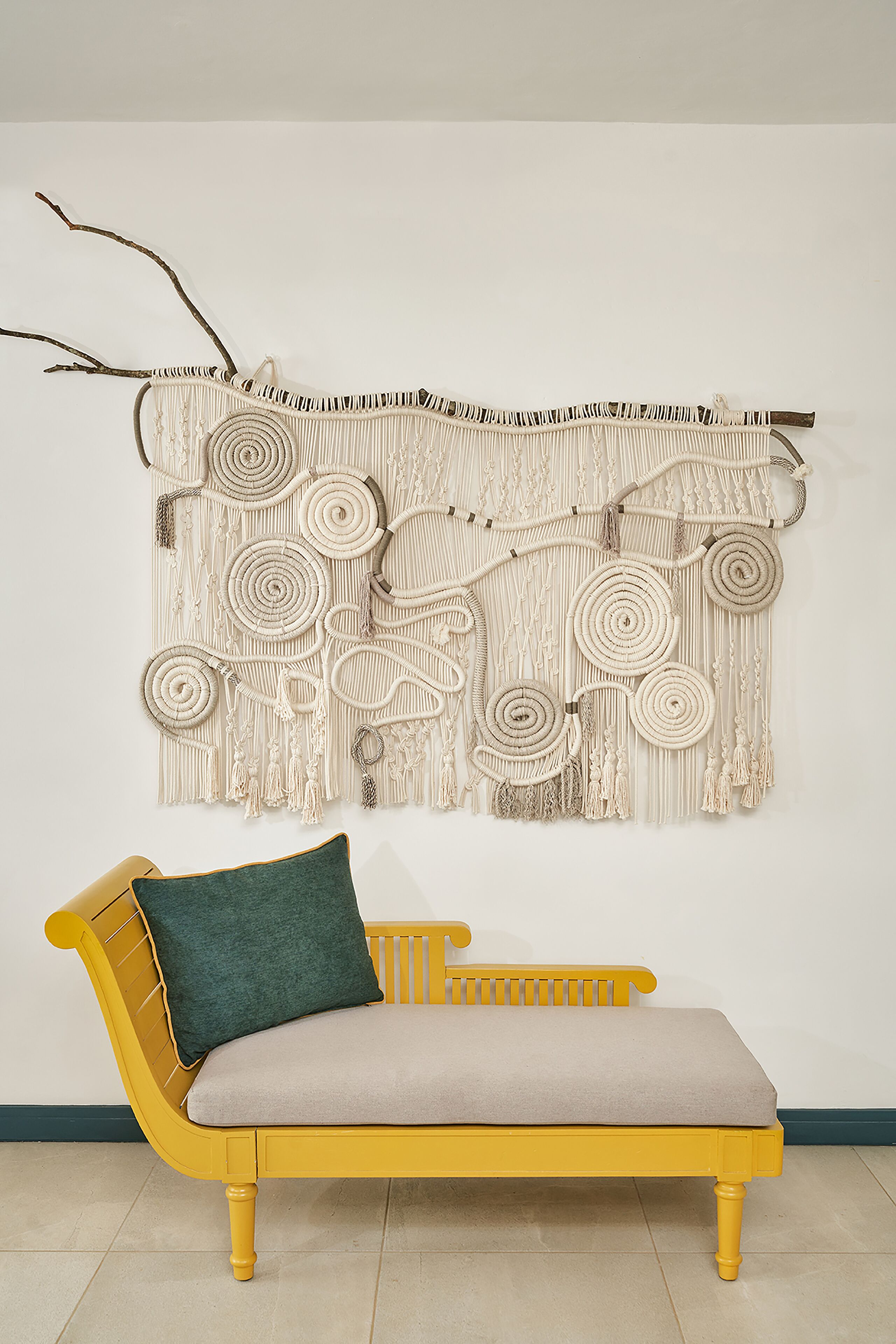 A yellow sofa with a green cushion and a macrame wall hanging above it.