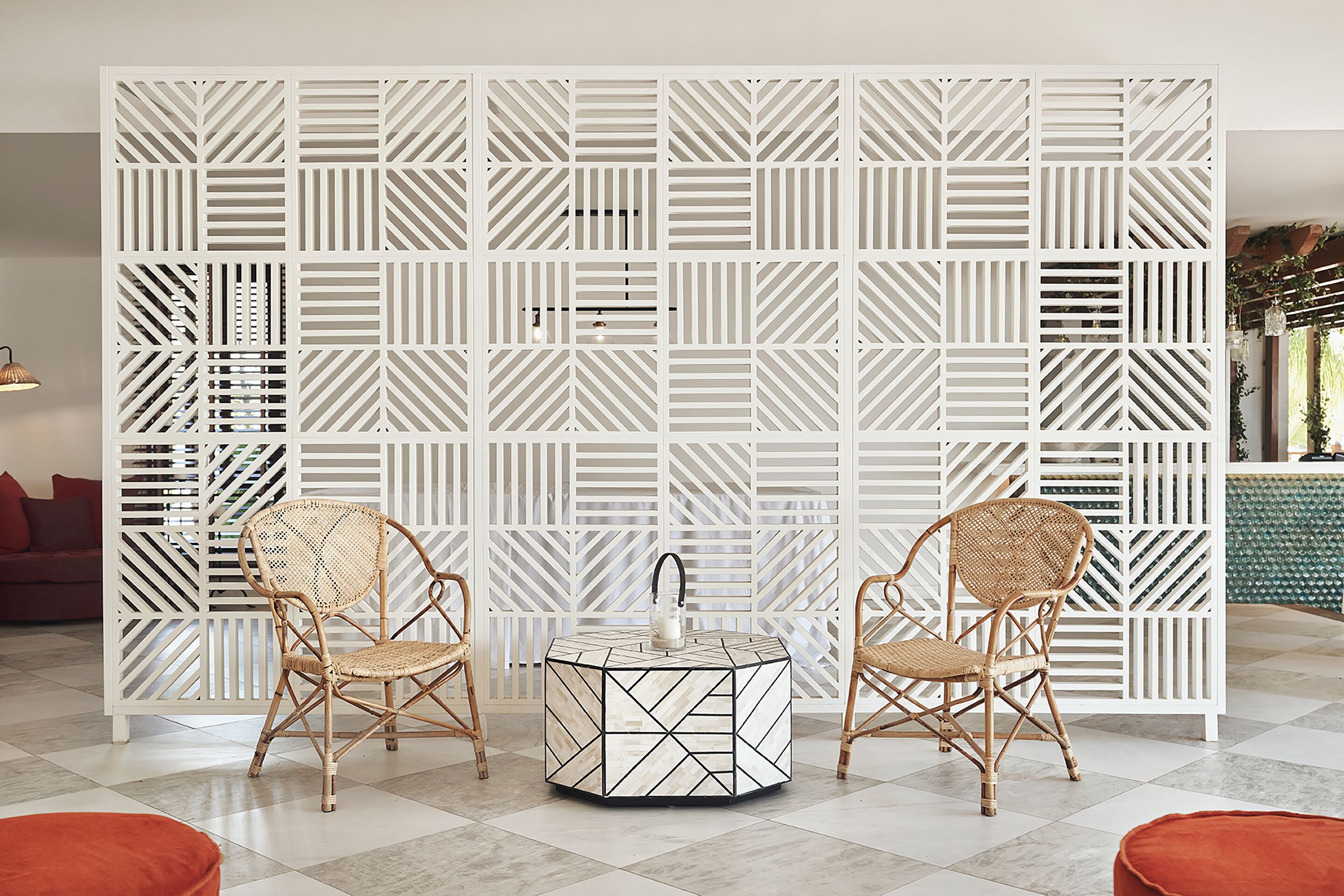 A room with a white geometric-patterned partition, two wicker chairs, a round table with a geometric design, and a red sofa.