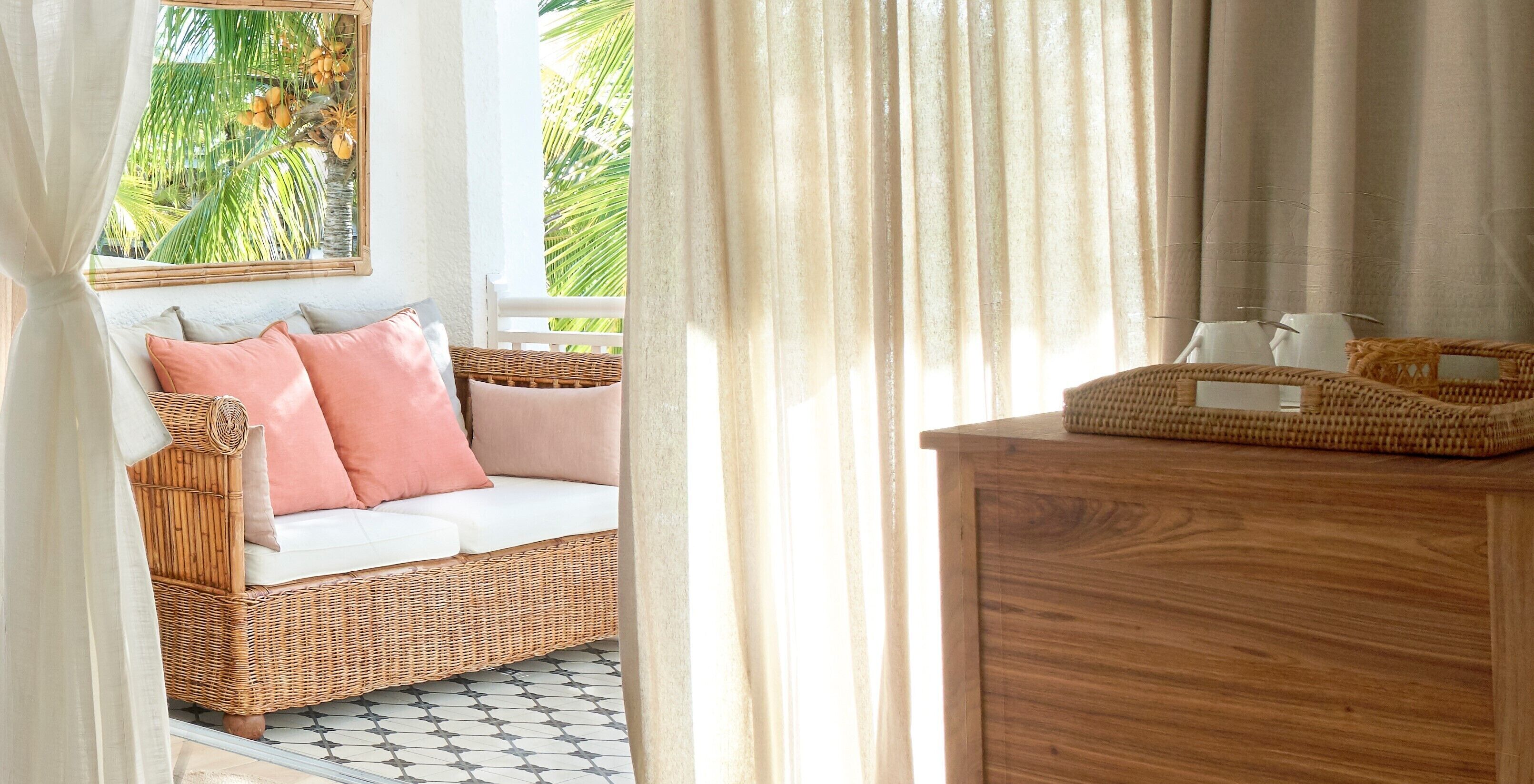 A wicker sofa with white and pink cushions, a wooden side table, and a view of palm trees through a window.