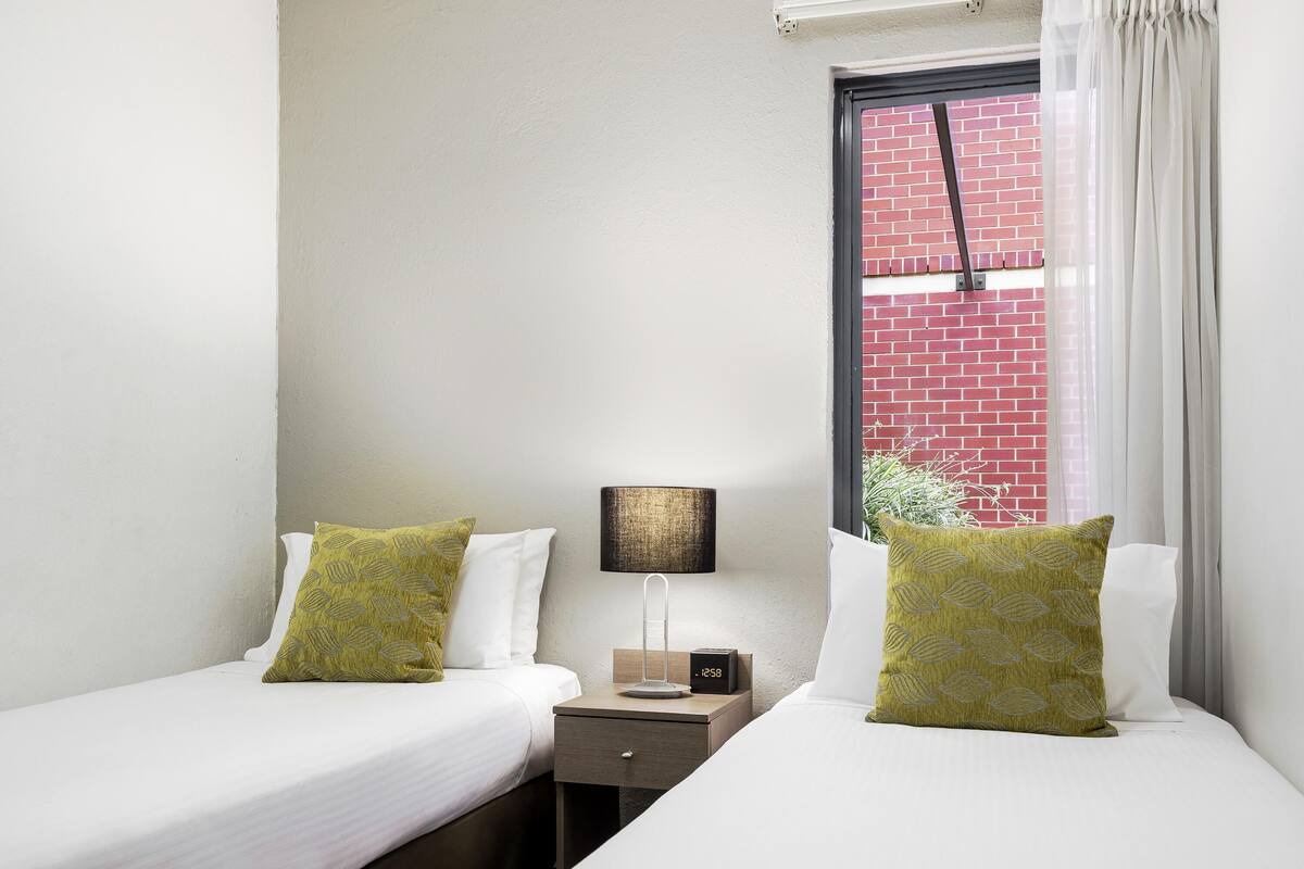 A twin room with two beds, a nightstand with a lamp, a clock, and a window with a view of a brick wall.