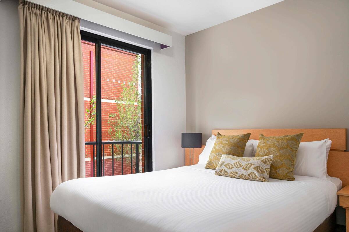 A hotel room with a large bed, a window with curtains, a wooden headboard, and a nightstand with a lamp.