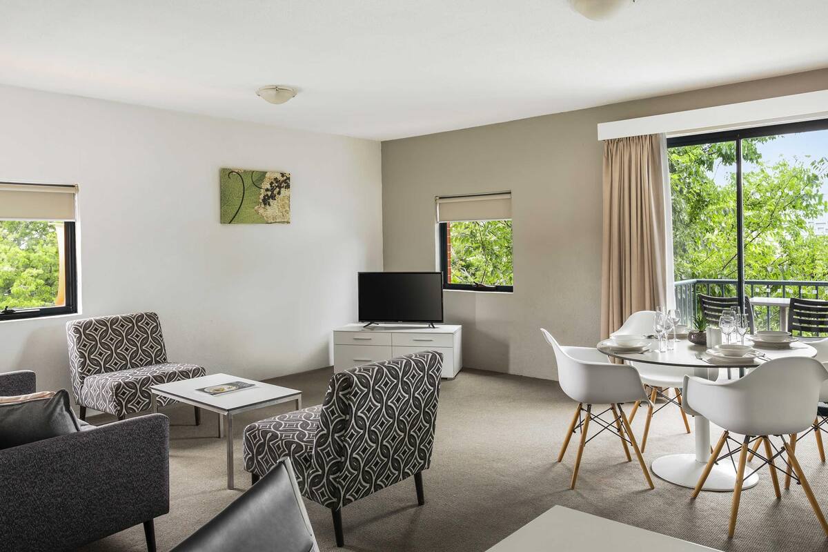 A modern hotel room with a flat-screen TV, a dining area with white chairs, a sofa, and a patterned armchair.