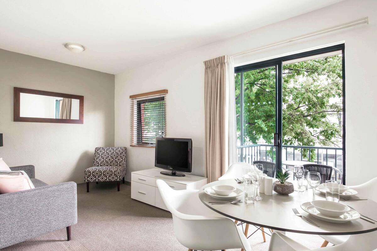 A modern living room with a sofa, a TV, a dining table set for two, and a balcony with a view of trees.