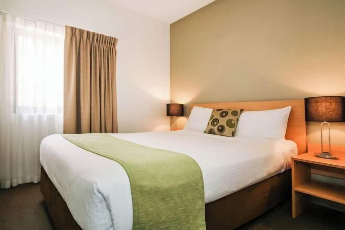 A hotel room with a bed, two bedside lamps, a nightstand, a window with curtains, and a wall-mounted lamp.