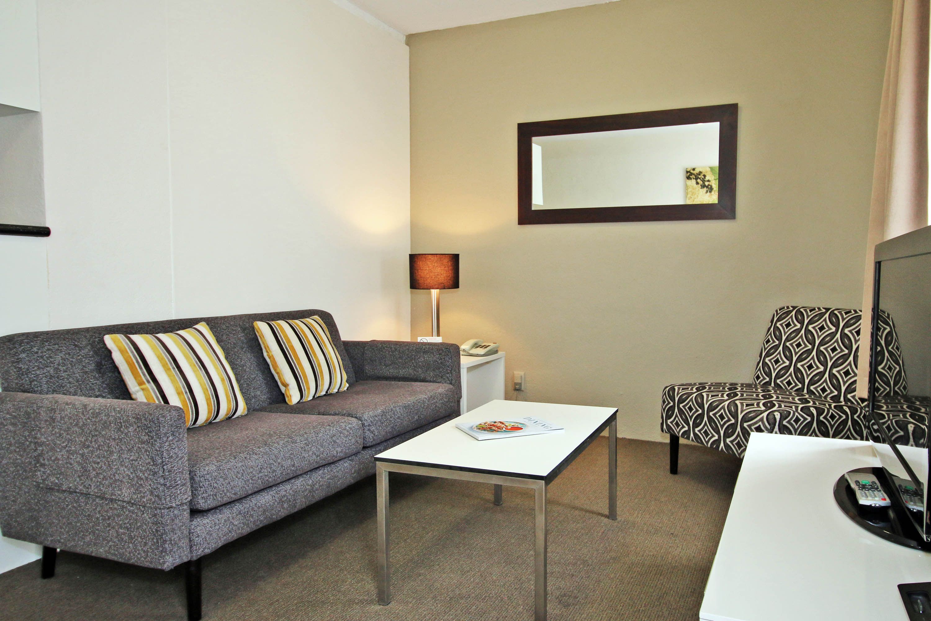A modern hotel room with a grey sofa, a white coffee table, a patterned armchair, and a small side table with a lamp.