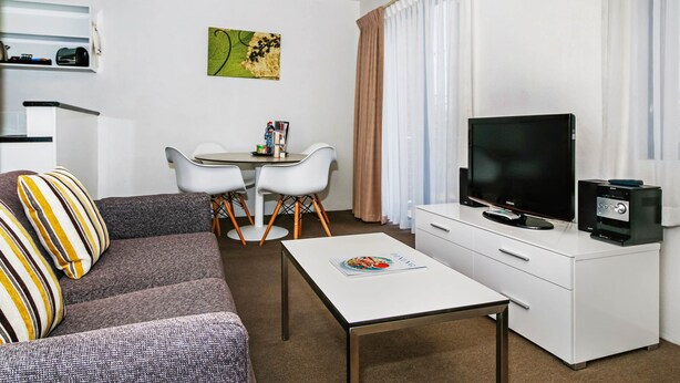 A modern hotel room with a gray sofa, a white coffee table, a TV on a white dresser, and a small dining table with chairs. - BreakFree Adelaide (Adelaide)