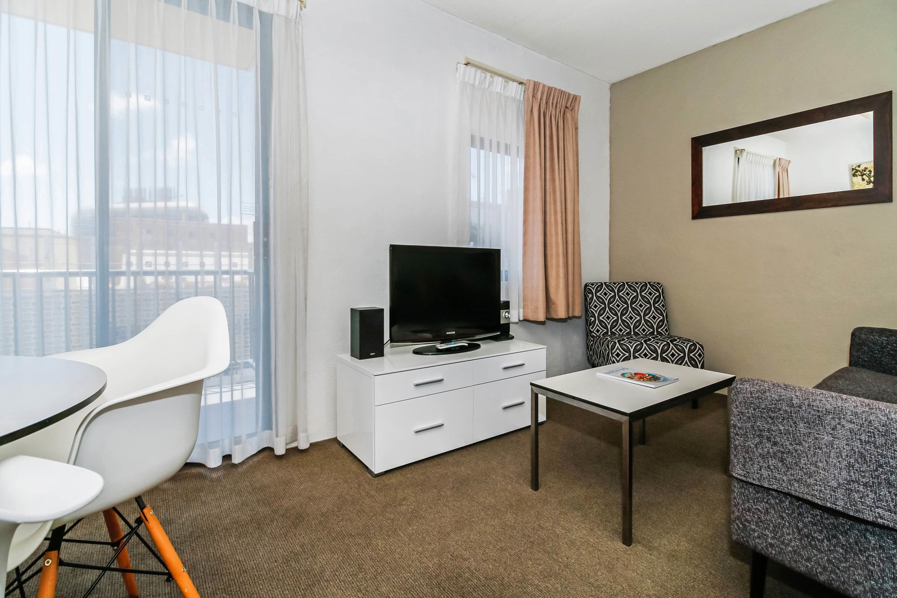 Habitación de hotel con un mueble blanco para televisor, un televisor de pantalla plana, una mesita, un sofá gris, una silla blanca y un sillón estampado.