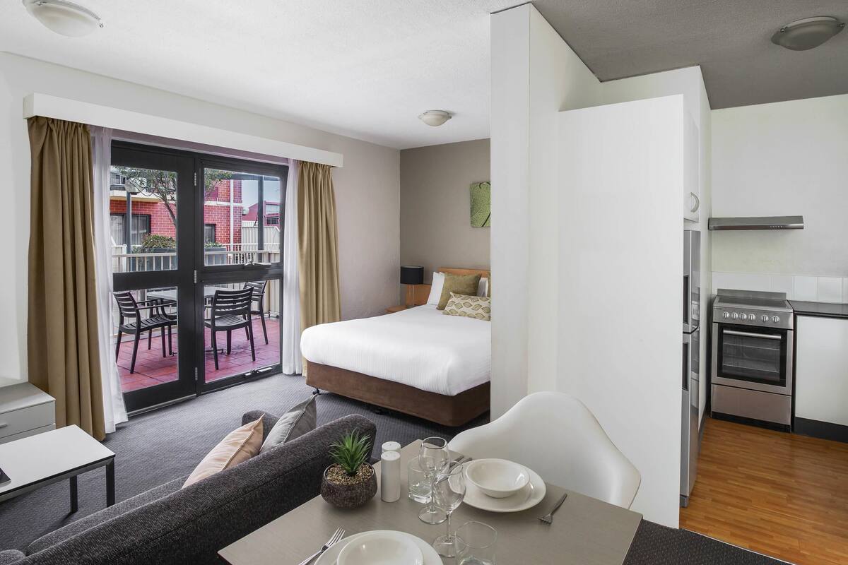 A hotel room with a bed, a dining table set for two, a kitchenette, and a balcony with a view of buildings.