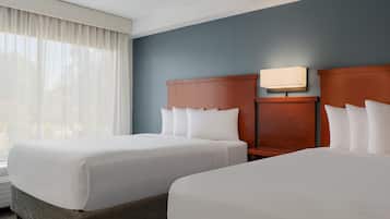 A hotel room with two beds, a nightstand, a wall-mounted lamp, and a large window with sheer curtains.