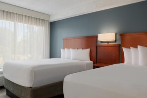 Habitación de hotel con dos camas, una mesita de noche, una lámpara de pared y un ventanal con cortinas translúcidas.