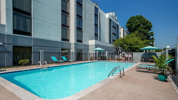 A swimming pool with lounge chairs and umbrellas, surrounded by a building with multiple windows and a tree. - Hyatt Select Nashville/Opryland (Nashville)