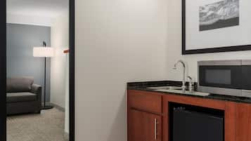 A compact hotel room with a kitchenette, a sofa, a floor lamp, and a framed artwork on the wall.
