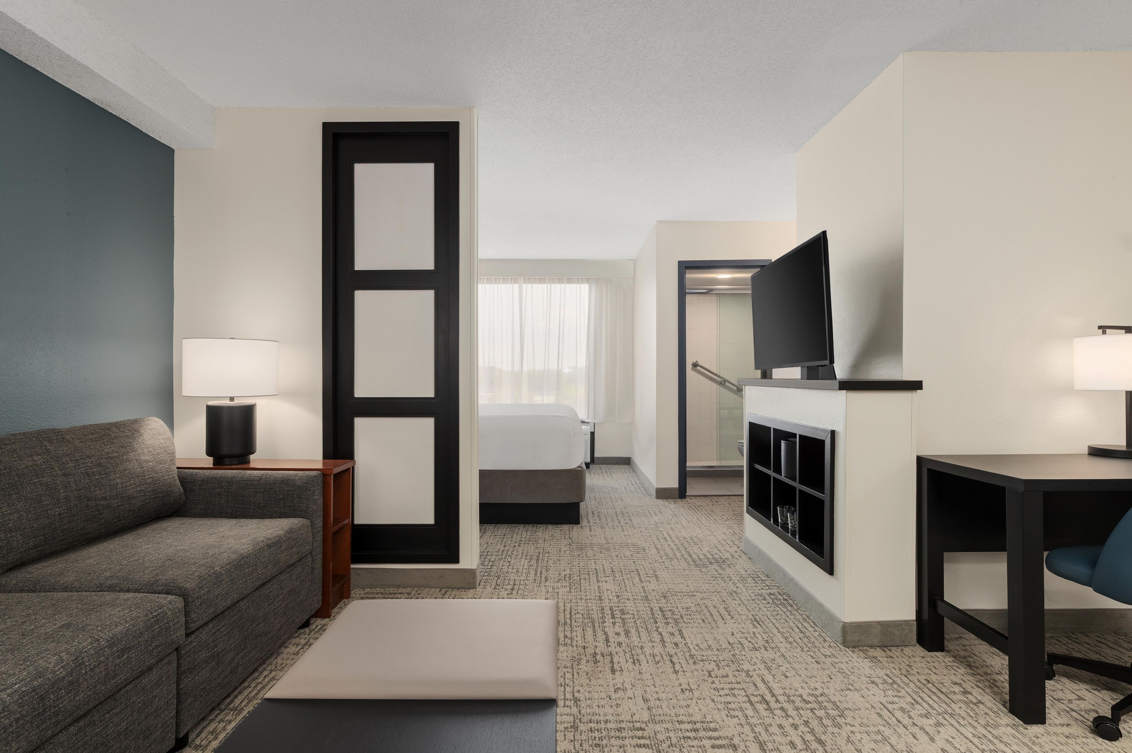 A hotel room with a sofa, a bed, a desk, a television, and a bathroom visible through an open door.