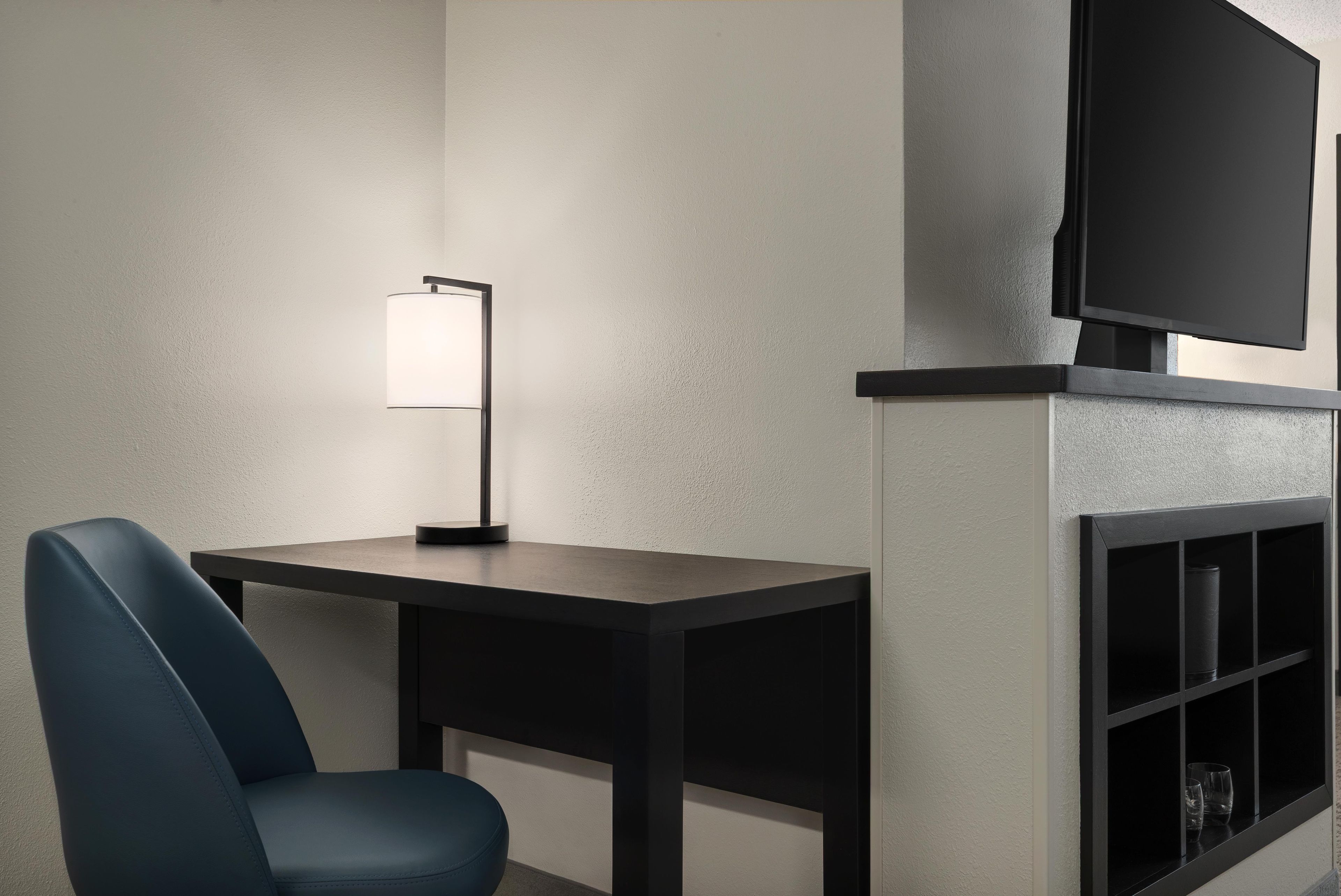 Una habitación moderna con un escritorio de madera oscura, una silla azul, una lámpara de pie y un televisor de pantalla plana montado en la pared.