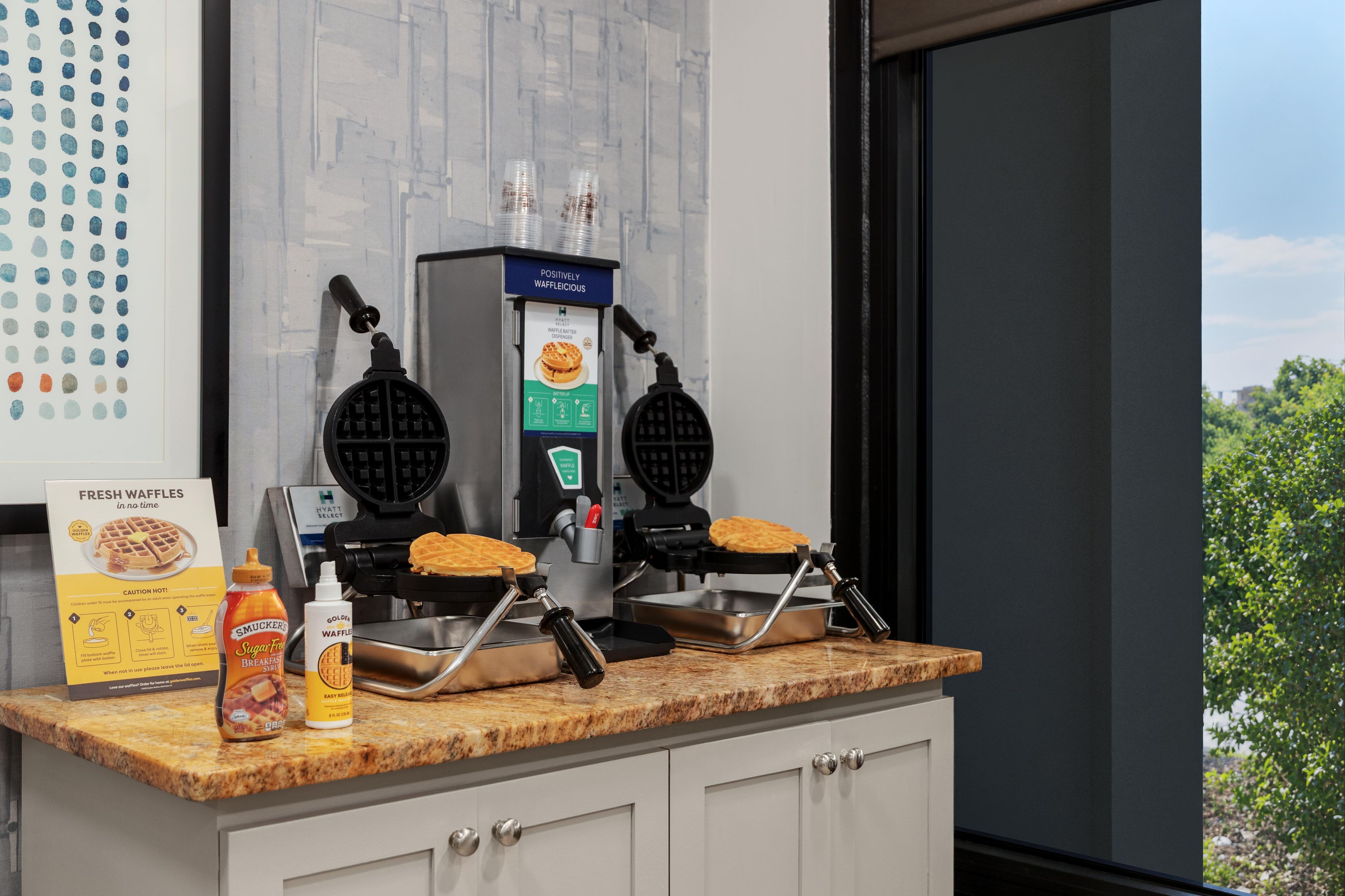 A kitchen area with two waffle makers, a box of fresh waffles, and a bottle of syrup.