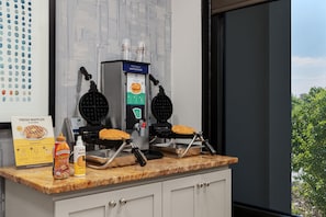 Un coin cuisine équipé de deux appareils à gaufres, d’une boîte de gaufres fraîches et d’une bouteille de sirop.