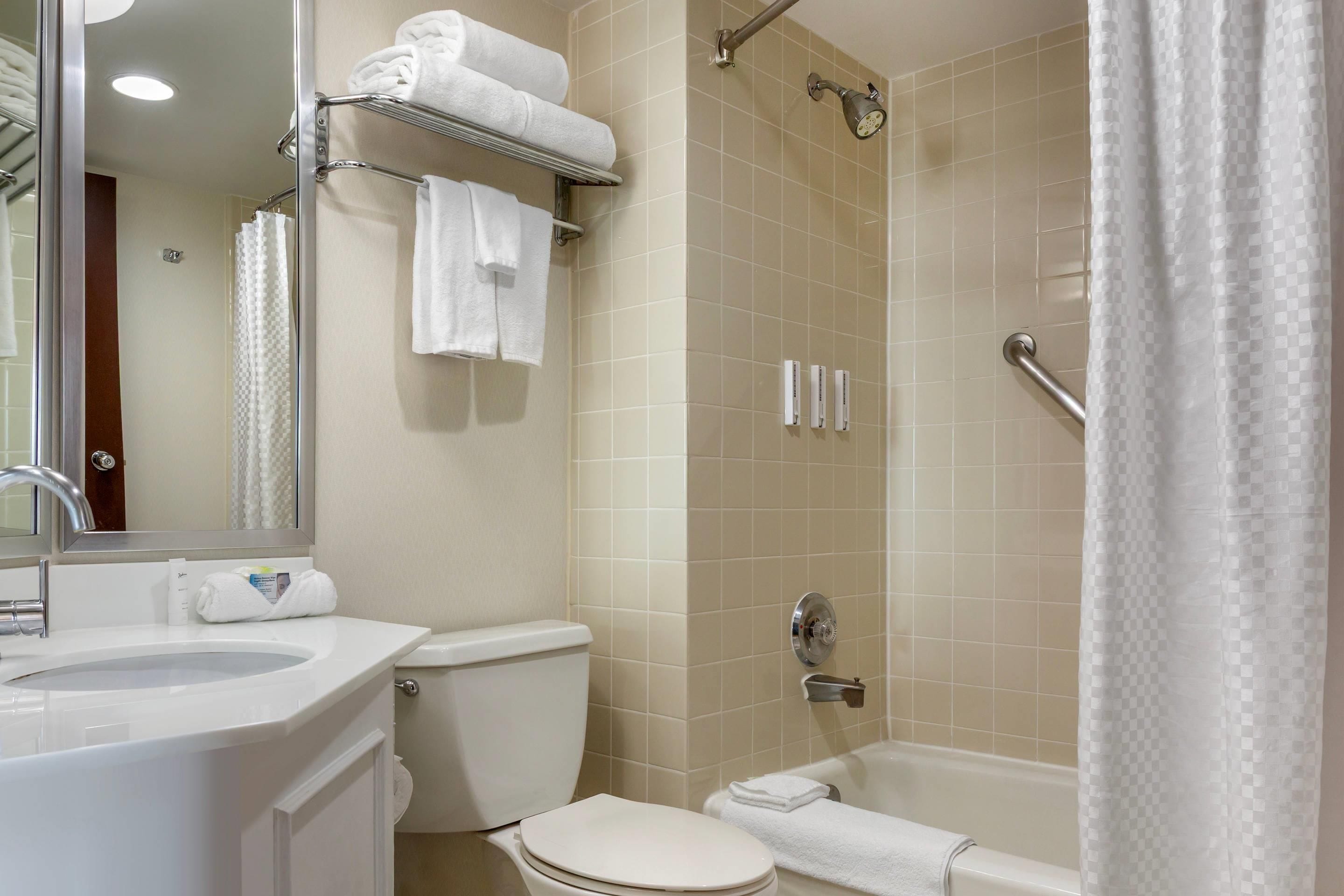 Baño con inodoro, lavabo y bañera. Hay un perchero con toallas, un cabezal de ducha y un pasamanos.