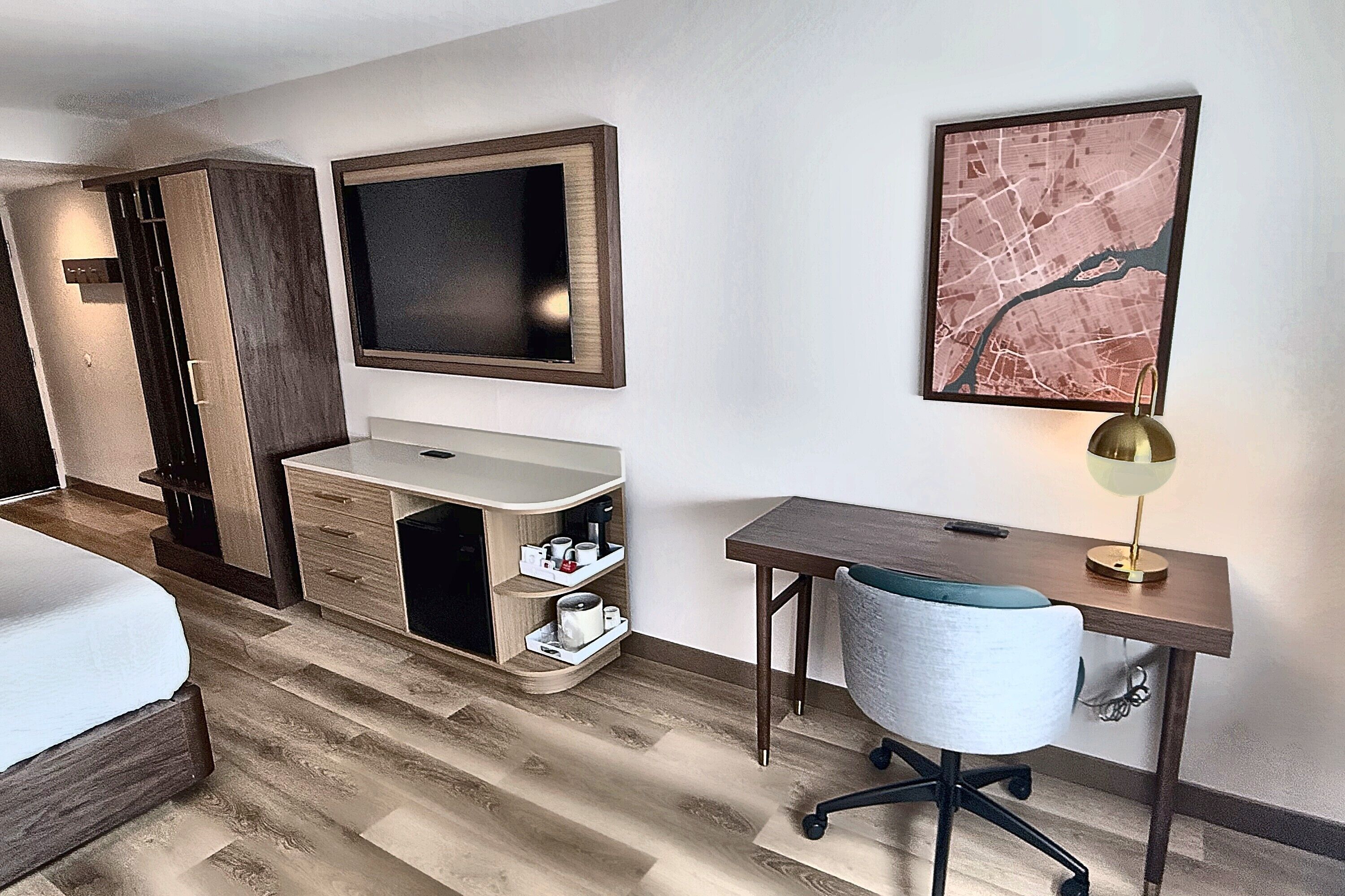A hotel room with a bed, a desk with a chair, a vanity with a mirror, and a framed map on the wall.