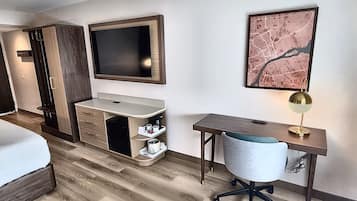A hotel room with a bed, a desk with a chair, a vanity with a mirror, and a framed map on the wall.