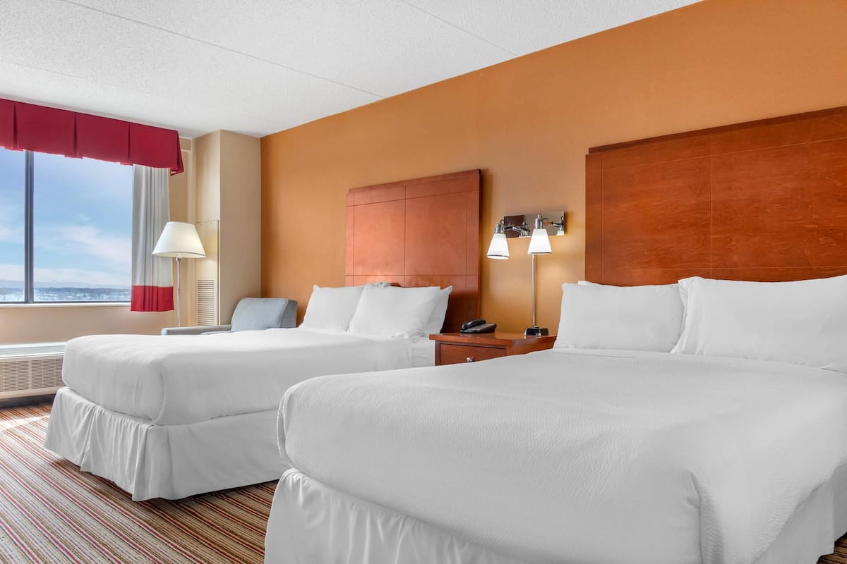 A hotel room with two beds, a wooden headboard, a nightstand with a lamp, and a view of the outdoors.