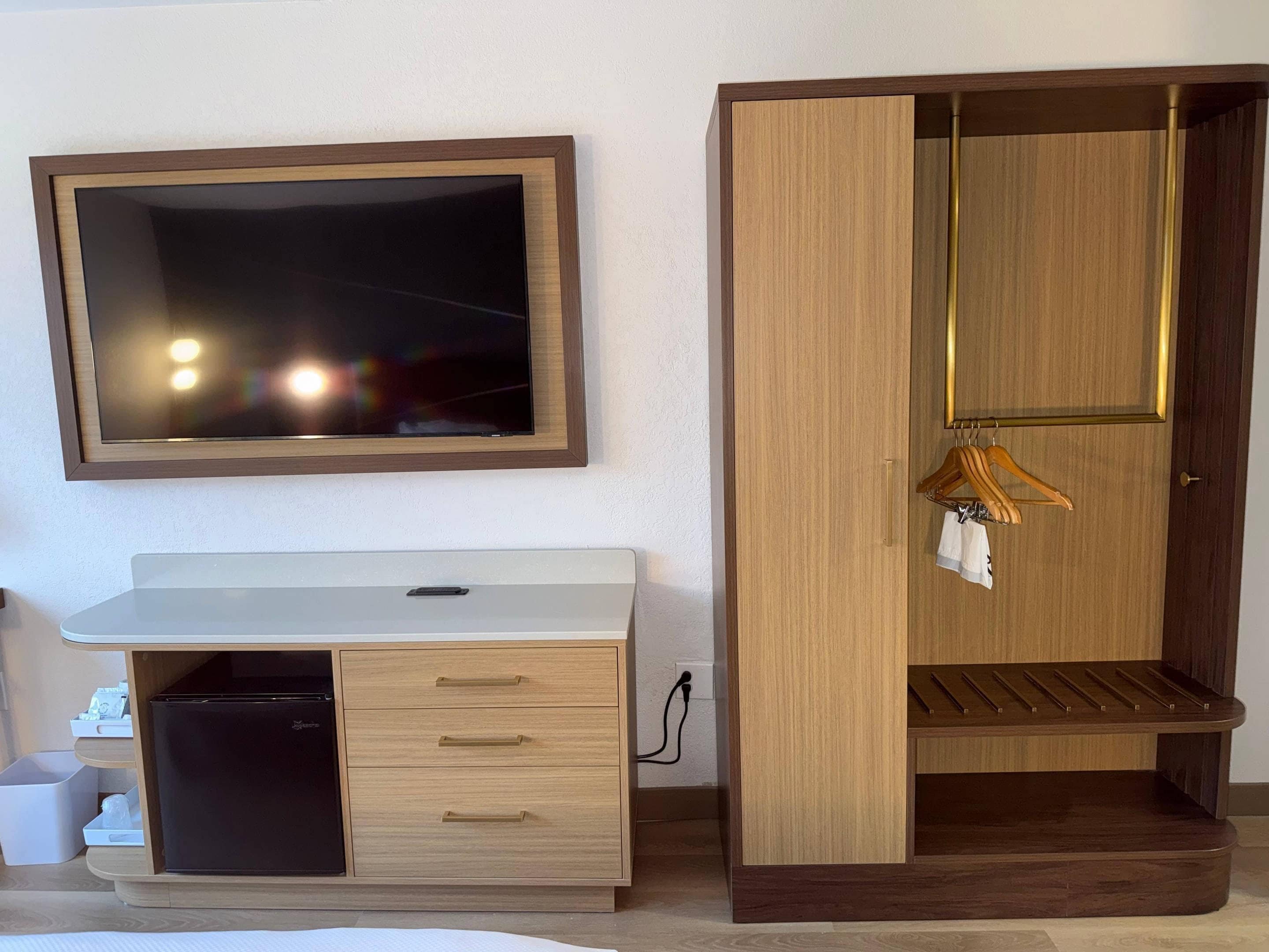 A room with a white desk, a black refrigerator, a wooden wardrobe with a mirror, and a hanging clothes rack.
