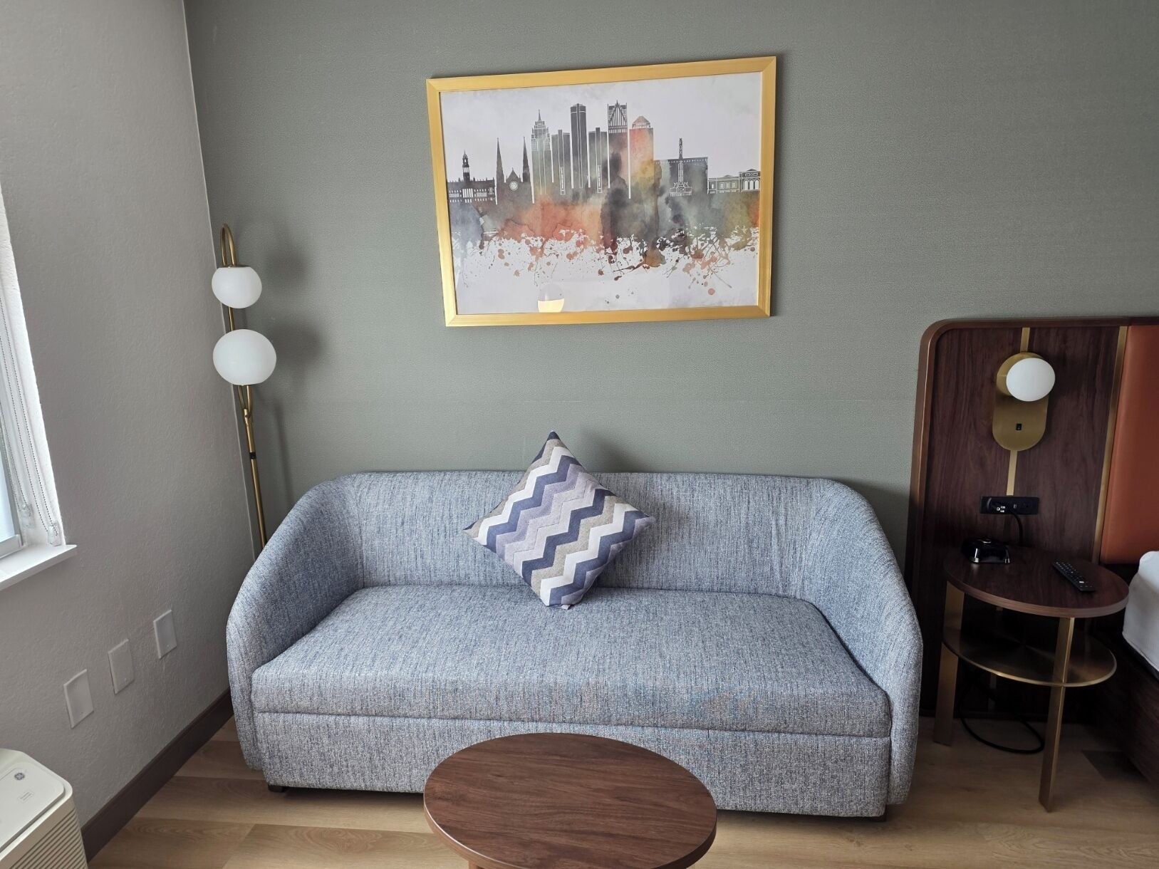 A modern living room with a grey sofa, a round wooden coffee table, a framed cityscape artwork, a floor lamp, and a bedside table with a lamp.