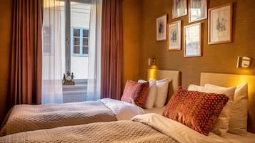 Habitación de hotel con dos camas, un ventanal con cortinas y cuadros enmarcados en la pared.