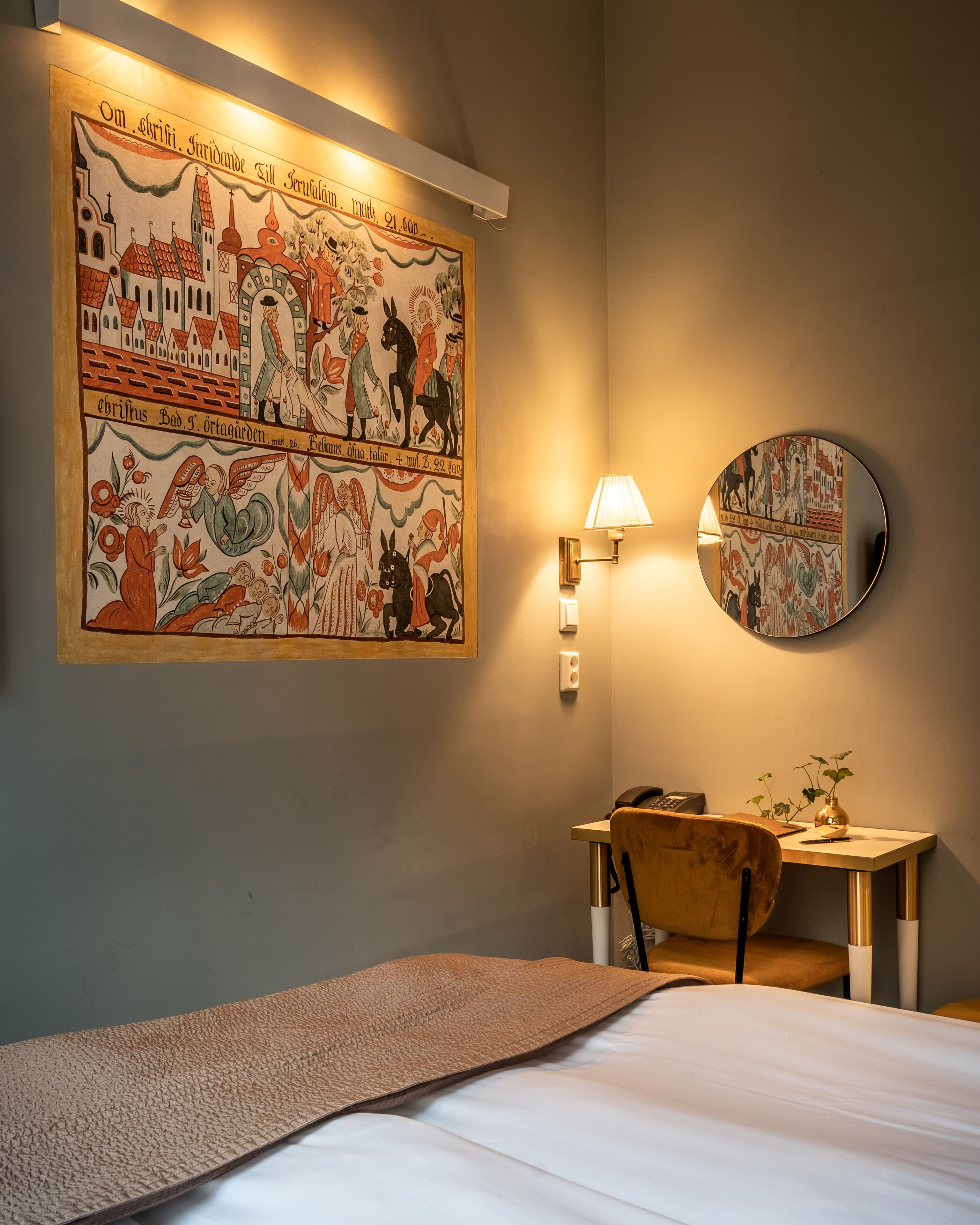 Quarto de hotel com cama, secretária com luminária, espelho fixado na parede e um grande mural retratando cenas históricas.