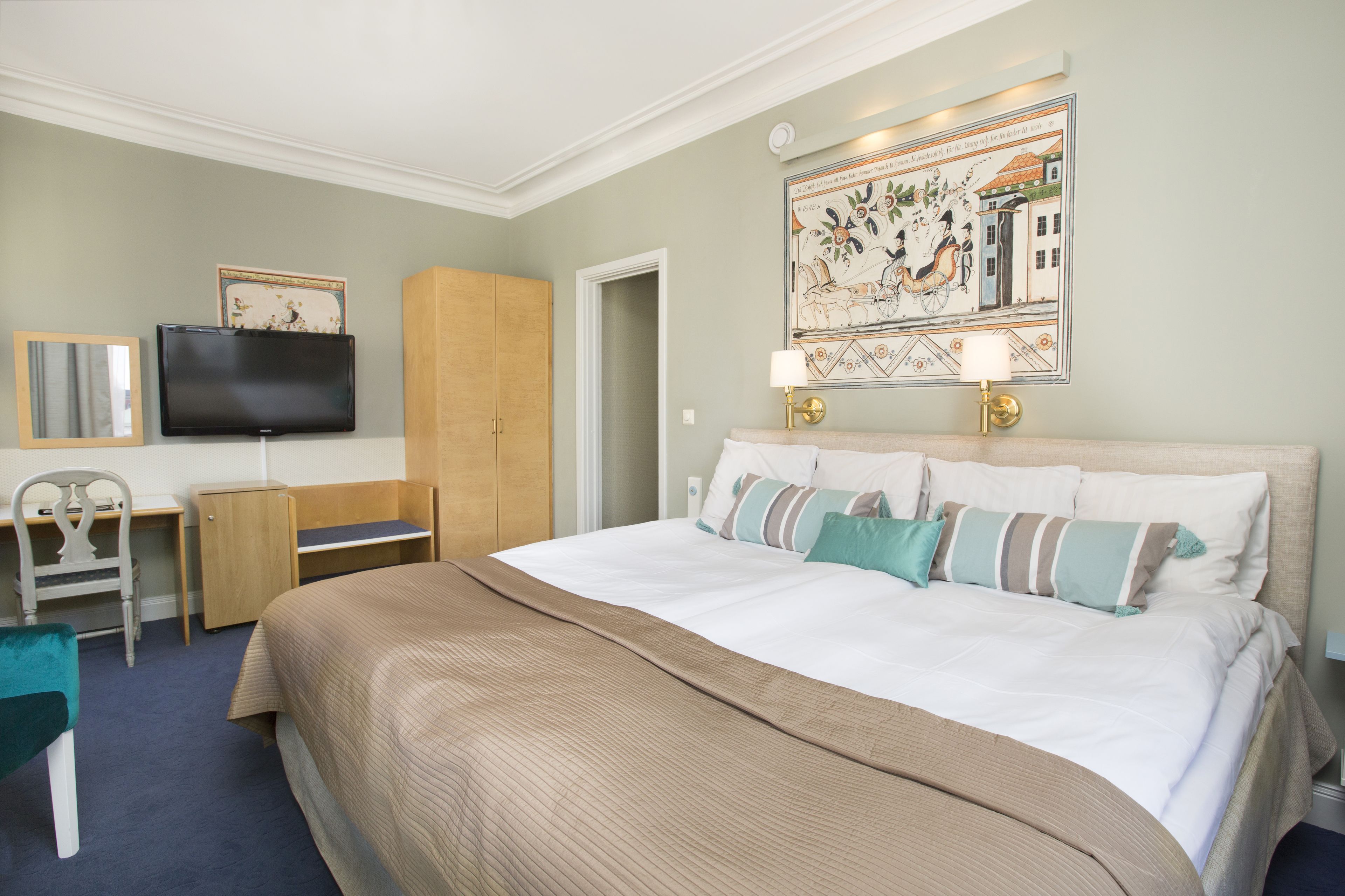Habitación de hotel con una cama grande, un escritorio, una silla, un televisor y una pared decorada con un cuadro.