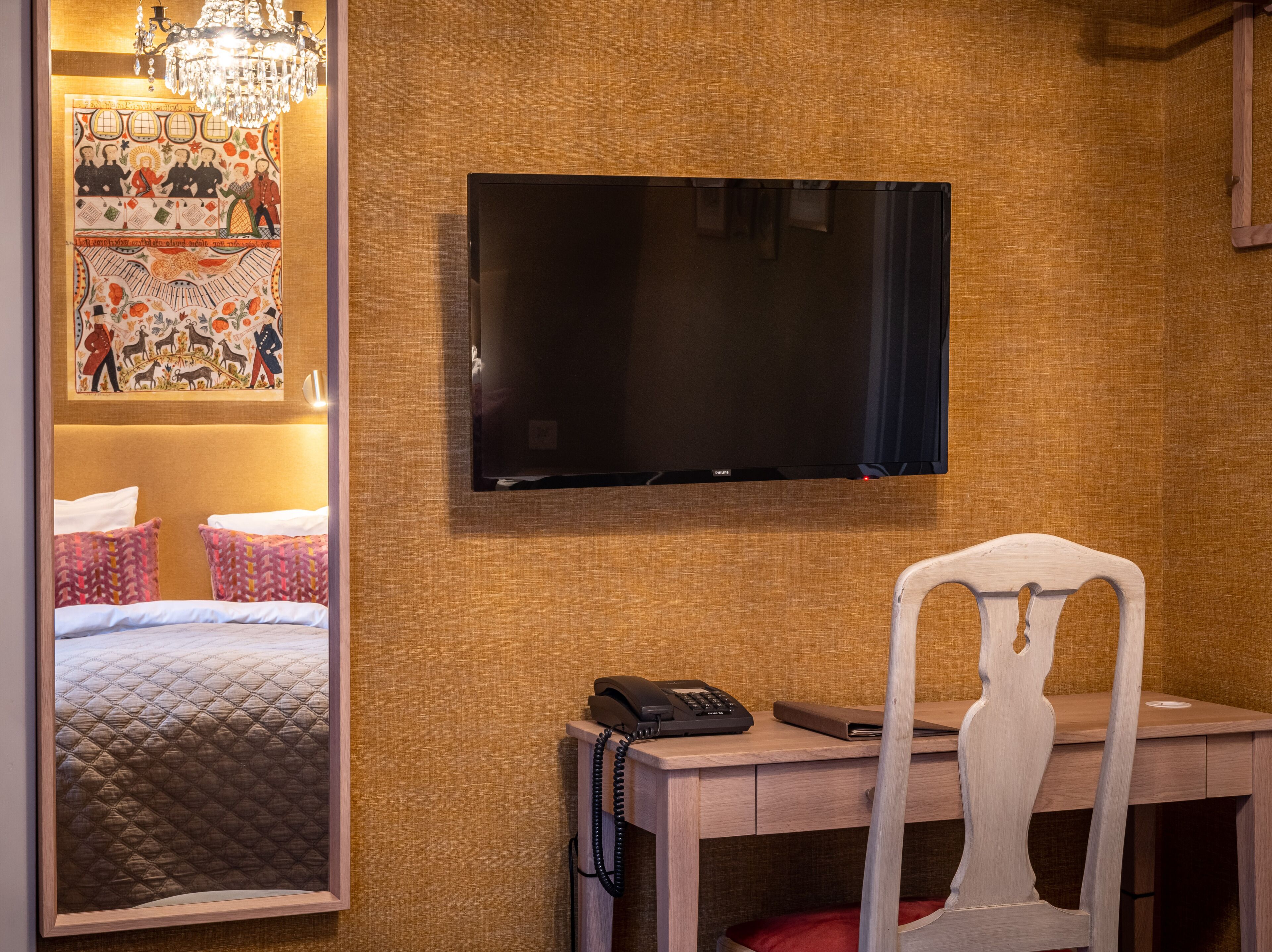 A hotel room with a bed, a desk with a telephone, a chair, a television mounted on the wall, and a decorative mirror.