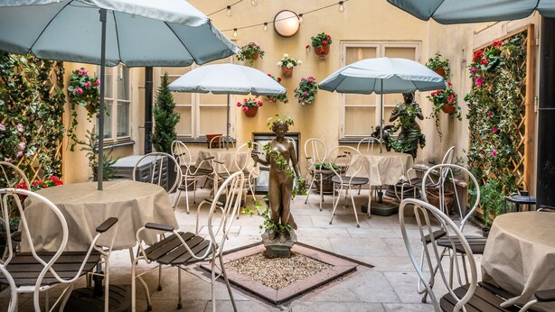 Un café al aire libre con mesas y sillas, cubierto por sombrillas y decorado con plantas en macetas y una estatua.