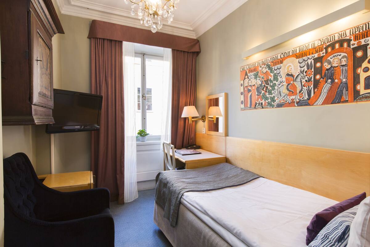 A hotel room with a bed, a desk, a chair, a television, and a painting on the wall.