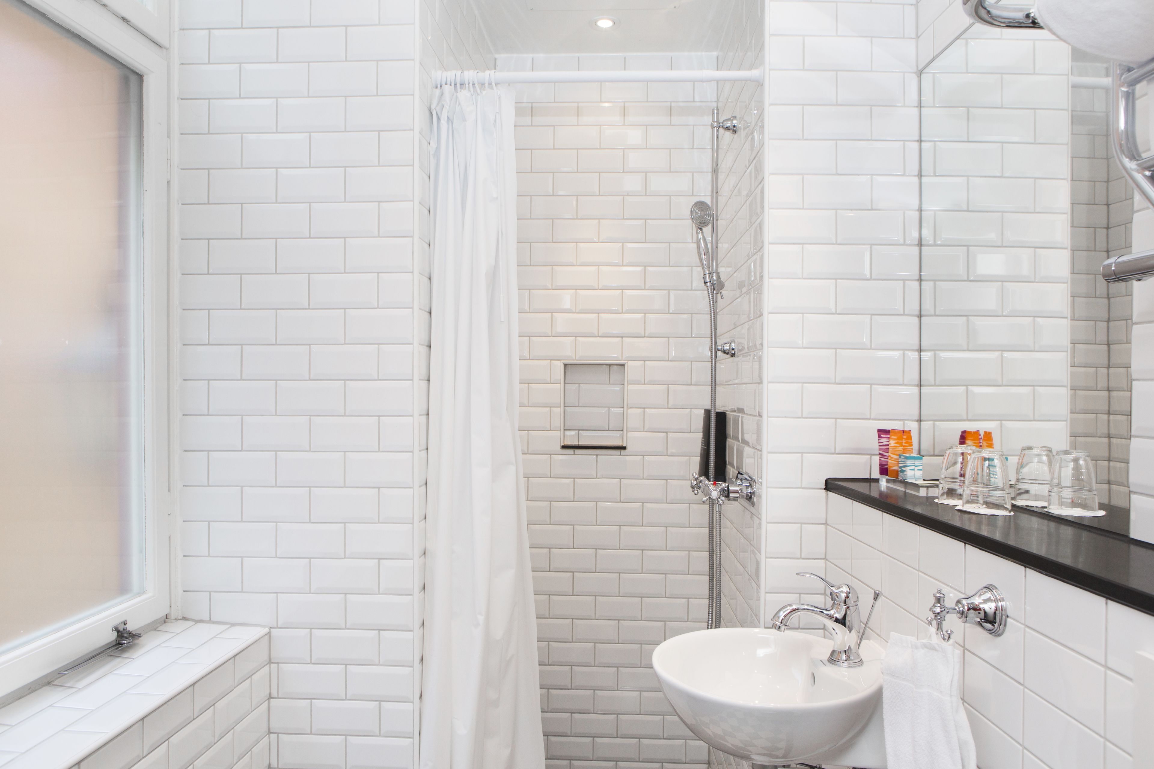 Een moderne badkamer met witte betegelde wanden, een glazen douchecabine, een witte wastafel en een zwart aanrechtblad met toiletartikelen.