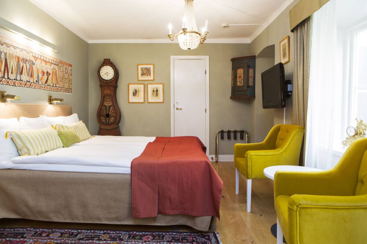 A hotel room with a bed, a desk, a chair, a television, a clock, and artwork on the walls.