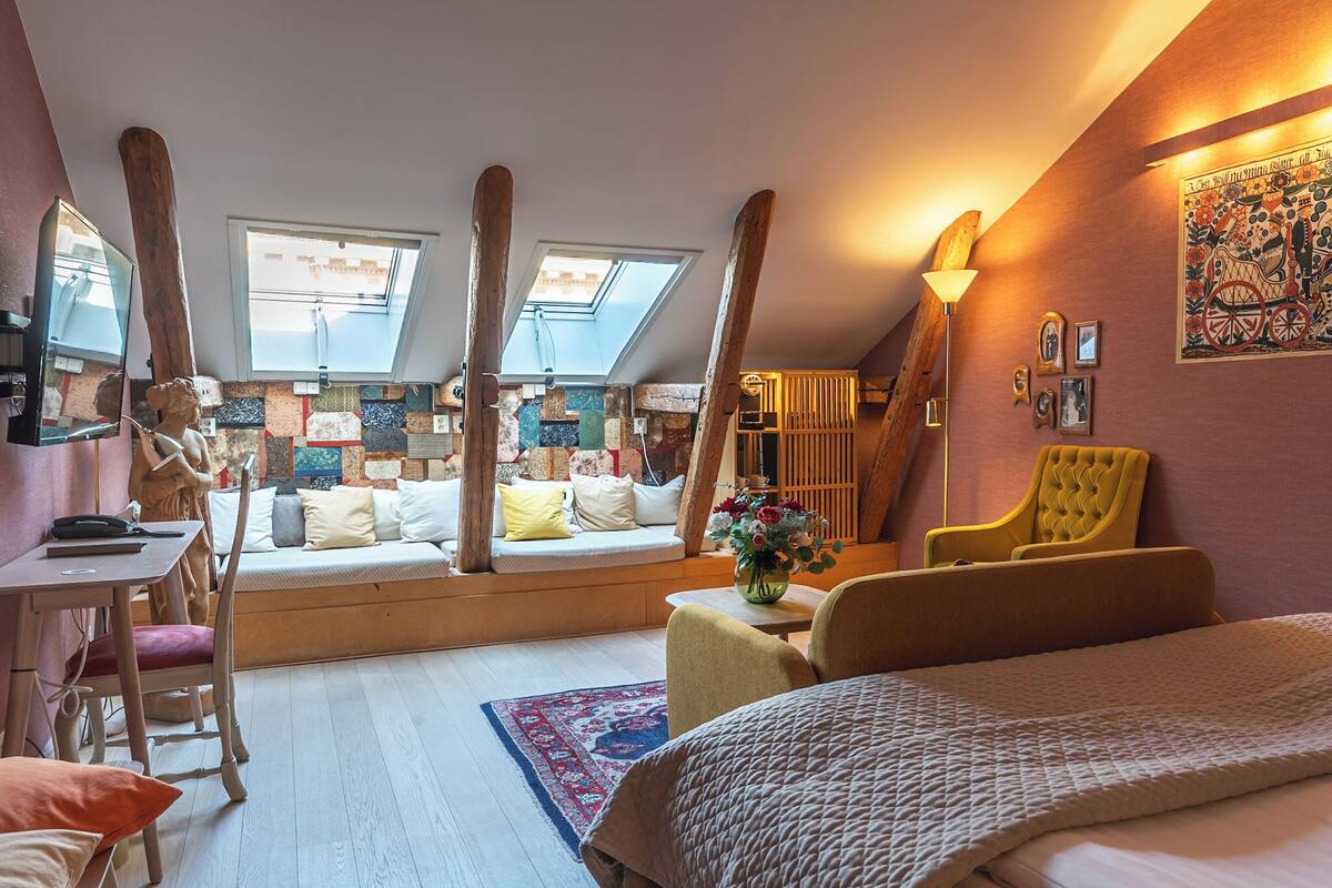 A cozy loft bedroom with a sloped ceiling, a large bed, a seating area with cushions, a small table with a vase, and a wooden chair.