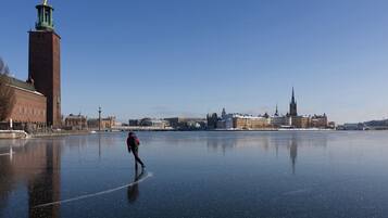 Un río congelado con una persona patinando sobre hielo, edificios al fondo y un cielo azul despejado.