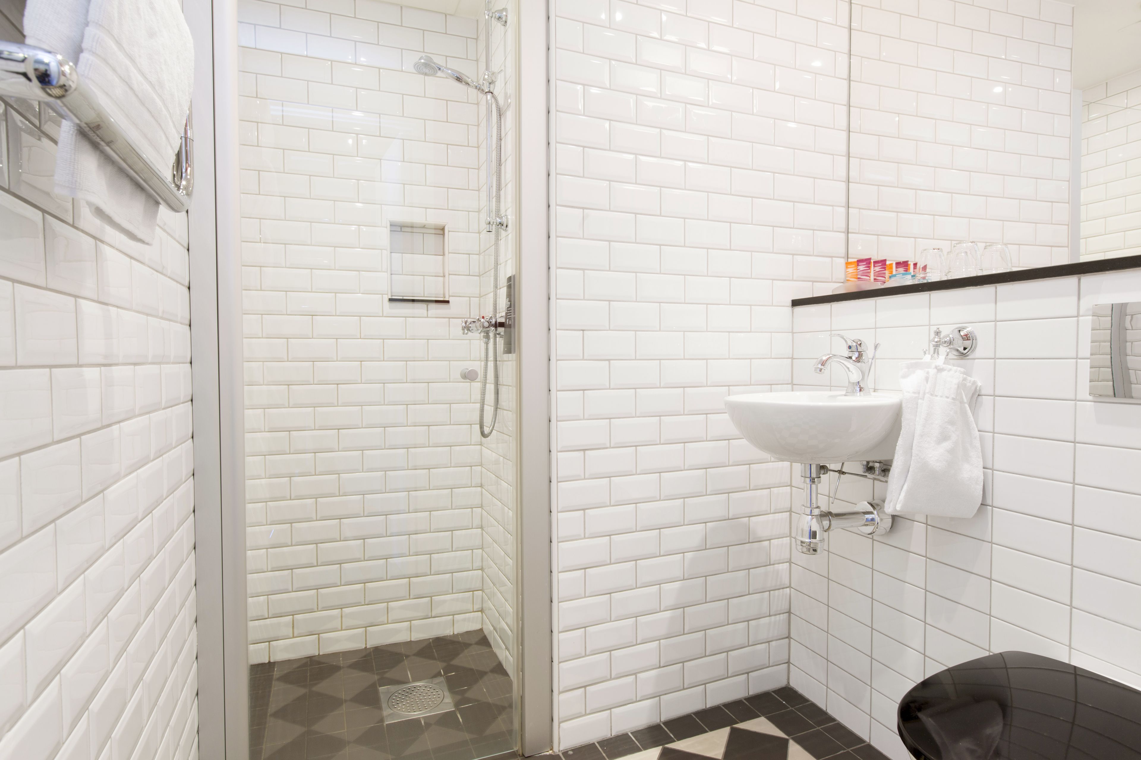 Un baño moderno con azulejos blancos tipo metro, ducha con cerramiento de vidrio, bañera negra y lavamanos blanco con una toalla colgada en la pared.