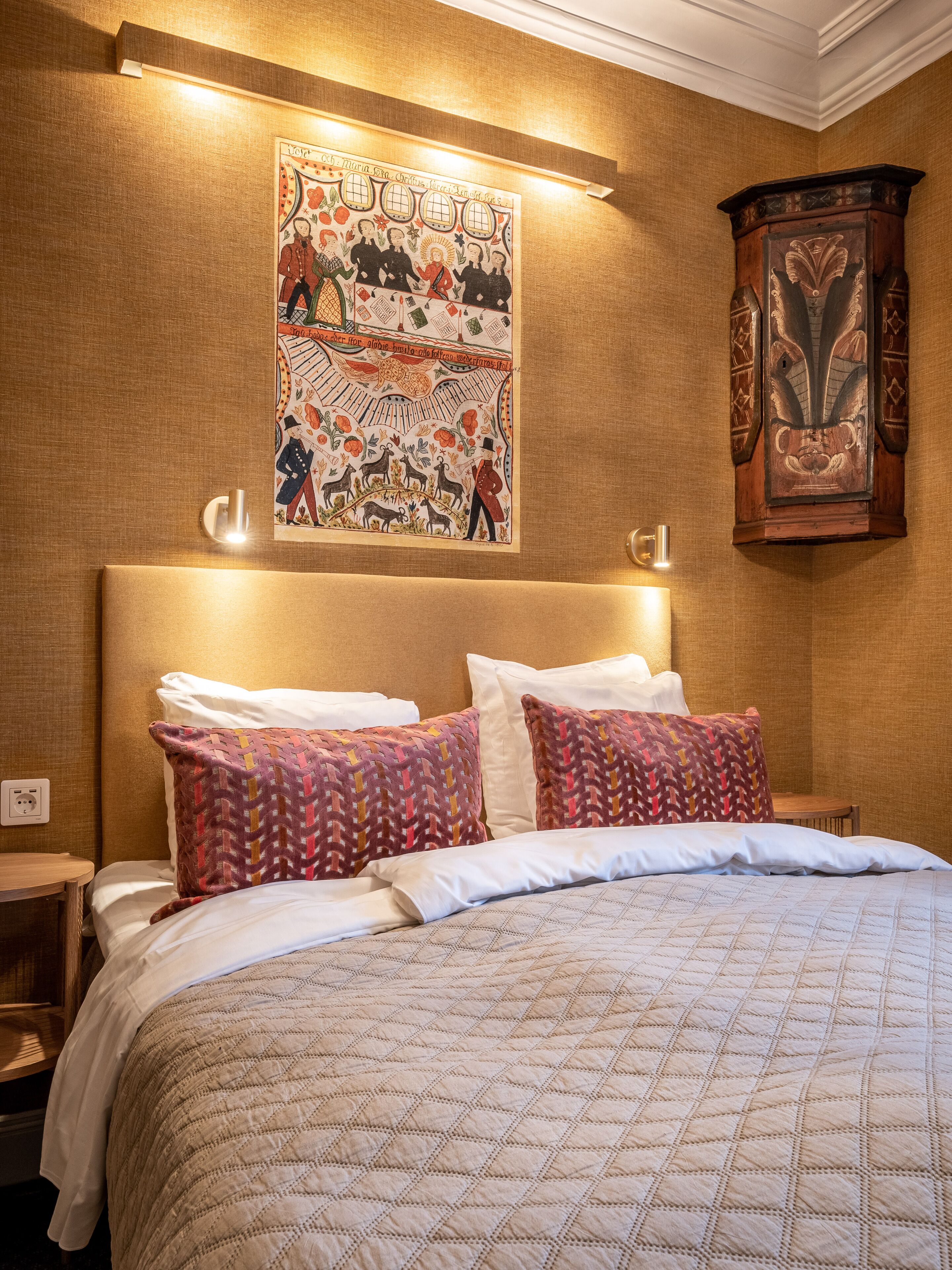 A neatly made bed with patterned pillows, a wall-mounted tapestry, and a wooden cabinet.