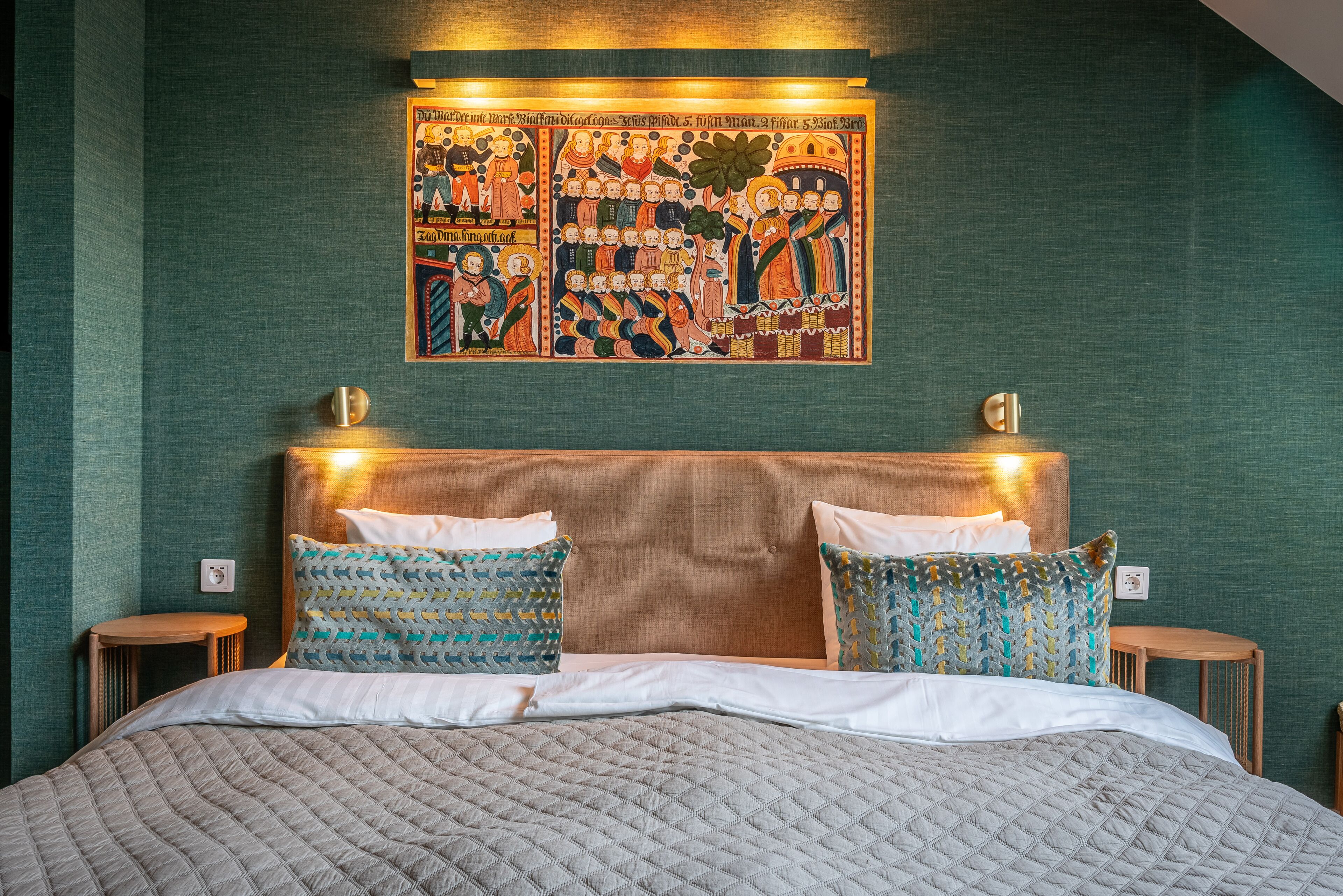 Un lit bien fait, agrémenté d’oreillers à motifs, une tête de lit avec des lampes fixées au mur, et une grande œuvre d’art à thème religieux au-dessus du lit.