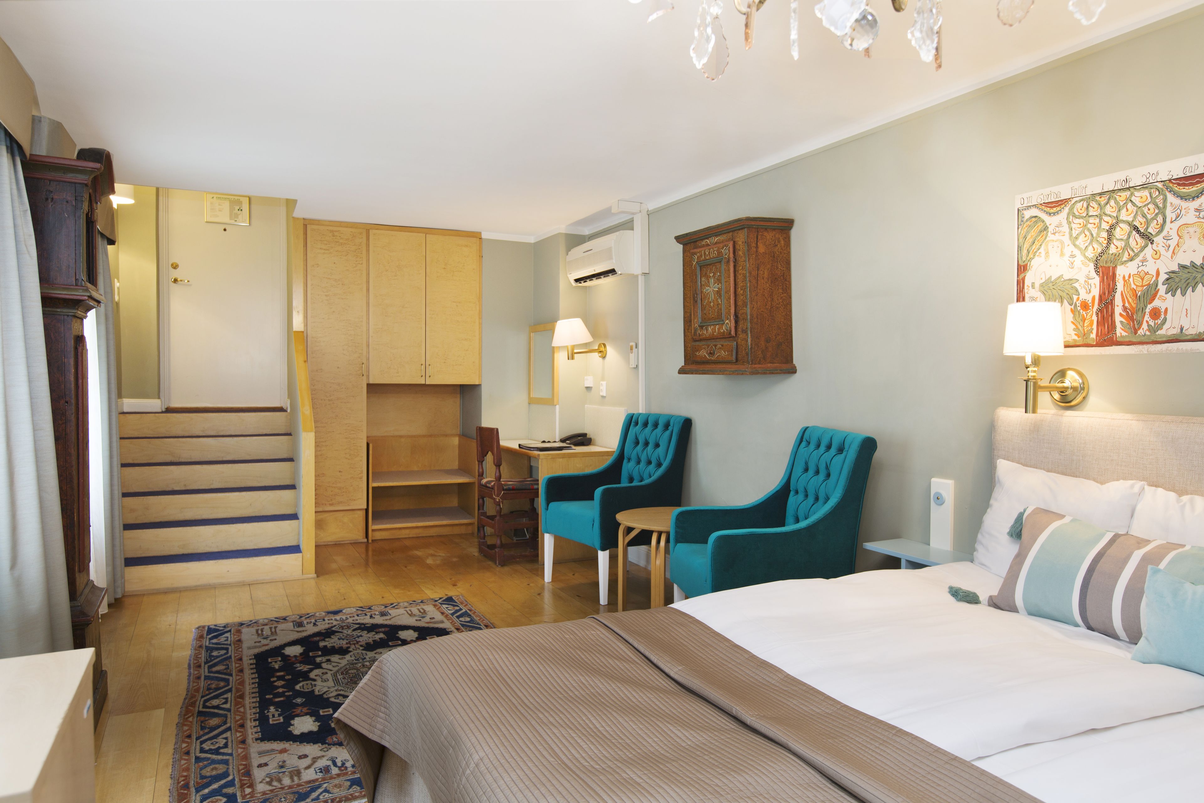 A hotel room with a bed, two chairs, a desk, a lamp, and a staircase leading to a closet.