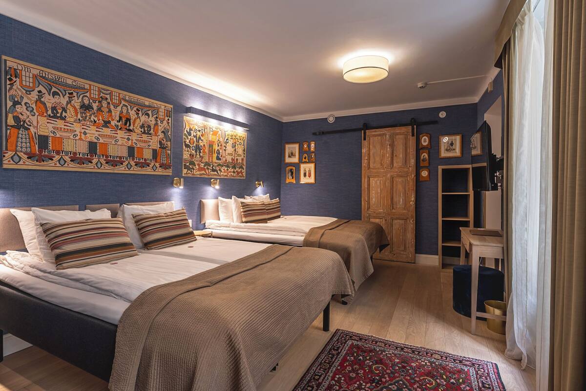 A hotel room with two beds, a large wall painting, a wooden wardrobe, and a small table with a chair.