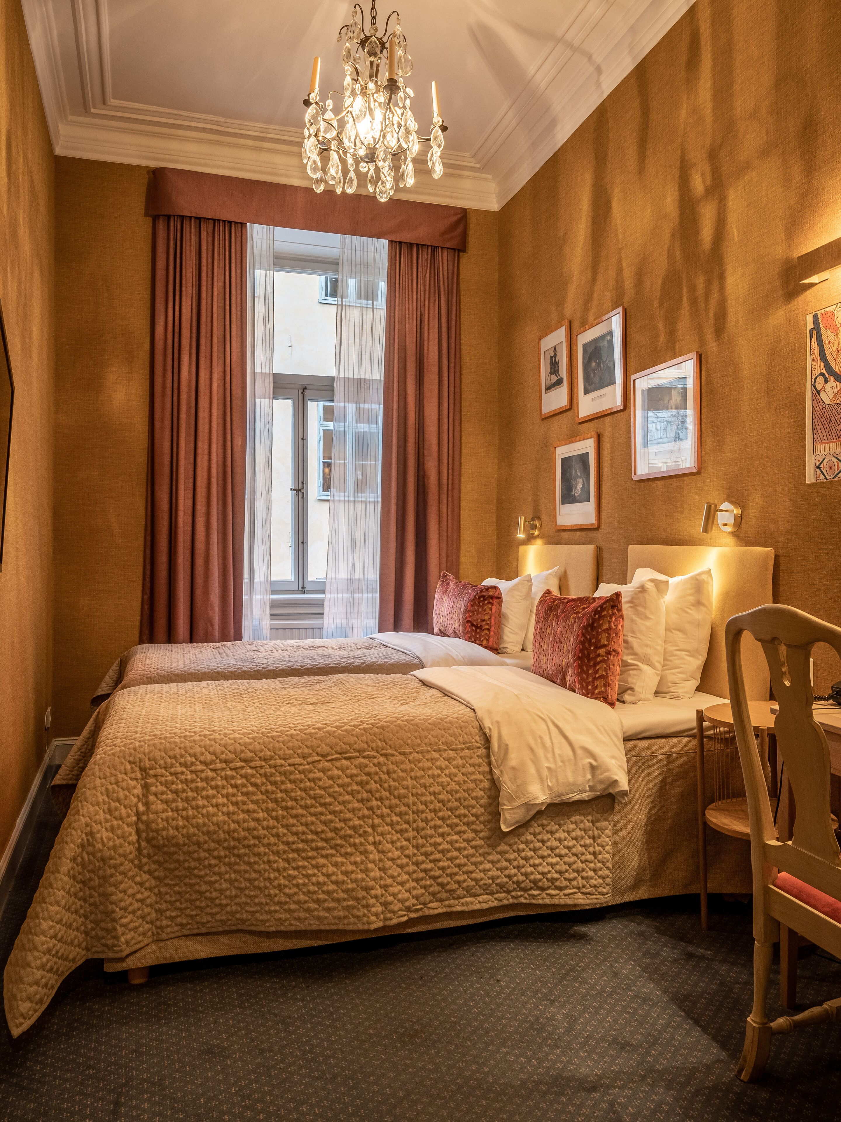 Ein Hotelzimmer mit zwei Betten, einer Chandelier, einem großen Fenster mit Vorhängen und gerahmten Bildern an der Wand.